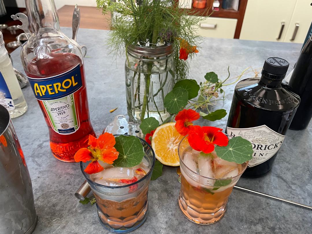 Two crystal glasses filled with a nasturtium spritzer and garnished with nasturtium flower and leaf. On the countertop next to the drinks is a bottle of Aperol, a bottle of Hendrik's Gin, a jar with water and fresh herbs, and silver cocktail shaker. 