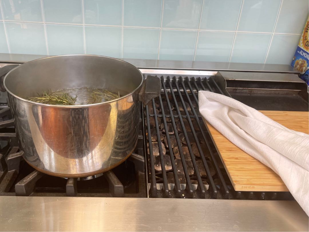 a pot of water on stove with rosemary sprig in it, small towel on counter next to the pot