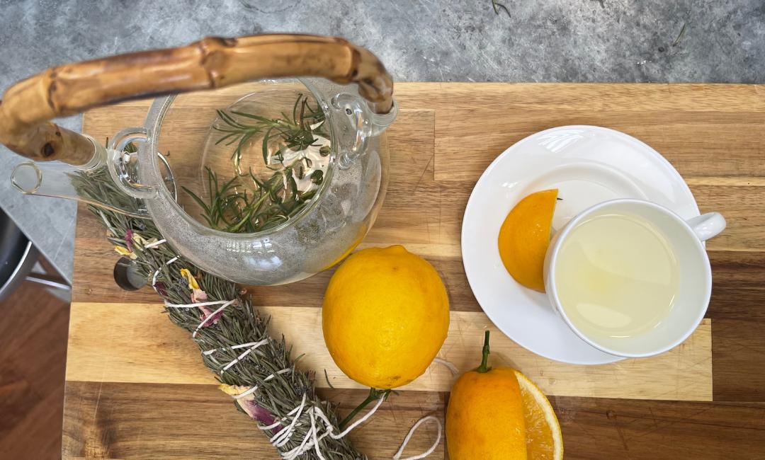 Rosemary tea in a glass teapot on wood cutting board next to rosemary smudge stick, lemons and cup of tea