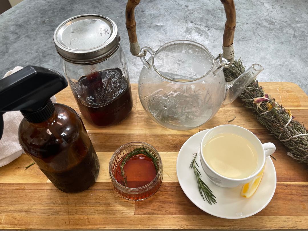 cutting board with a spray bottle, a jar of rosemary water, a cup of tea with lemon, a teapot, and some rosemary
