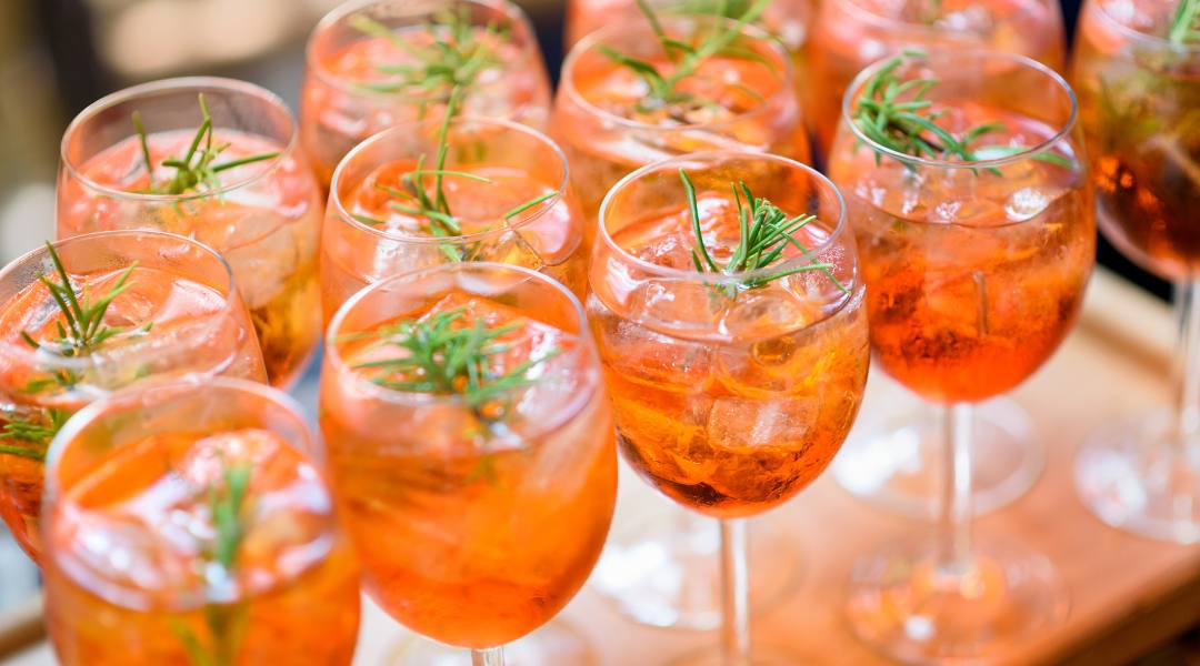 rows of wine glasses with an orange-toned cocktail, all garnished with fresh herbs