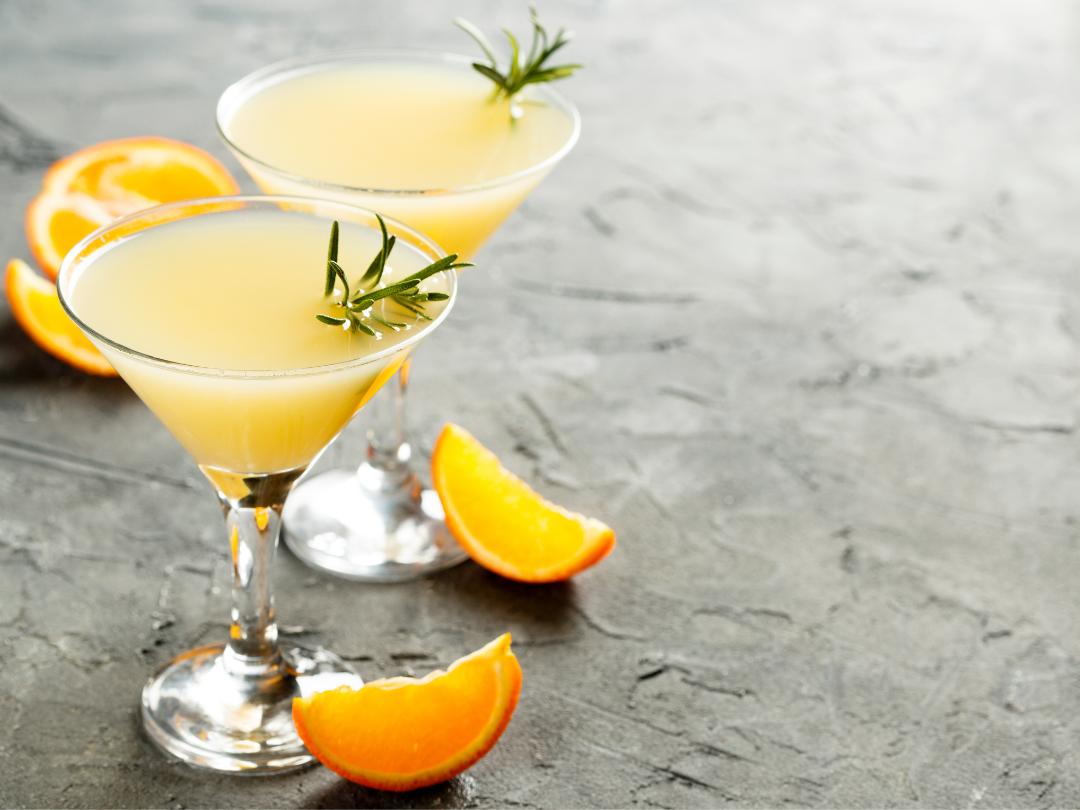 two frosty orange hued cocktails in martini glasses, garnished with a sprig of fresh rosemary