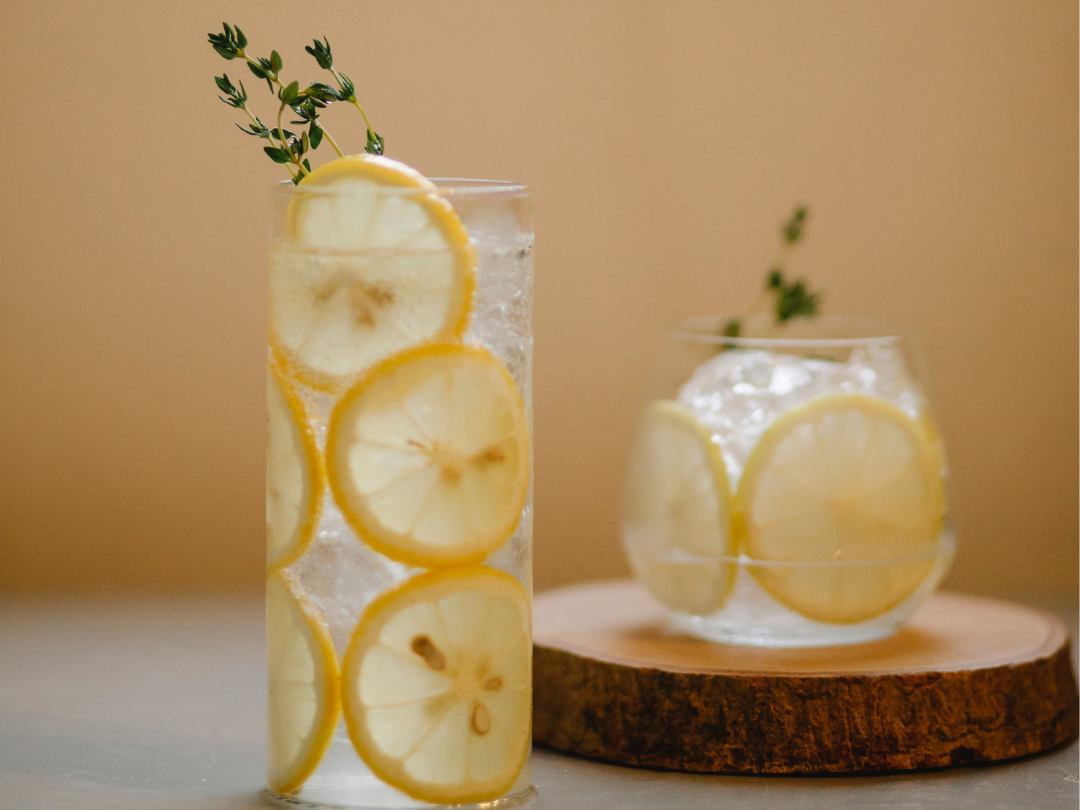 one tall narrow glass and one round glass, both filled with vodka lemonade. lemon slices and garnished with a sprig of fresh thyme